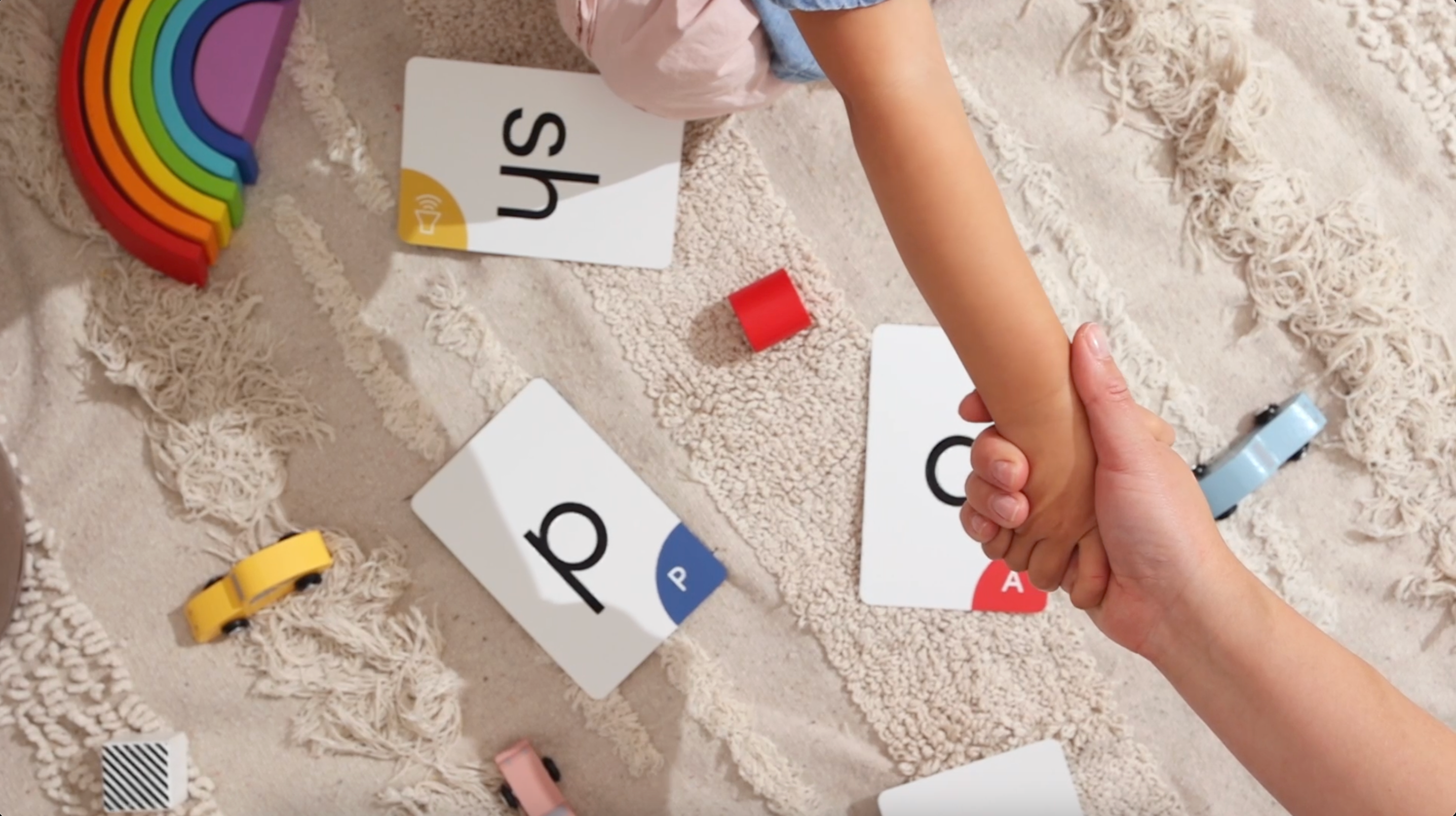 A small child hand and arm is shown, their hand is grasping a larger adult hand, squeezing it. Below is a cream color texture rug, with some toys scattered around including a bright rainbow made out of wood, some small wooden cars in yellow, pink and blue, some small wooden blocks and 3 alphabites game cards with the letters sh, p, and a written on them.