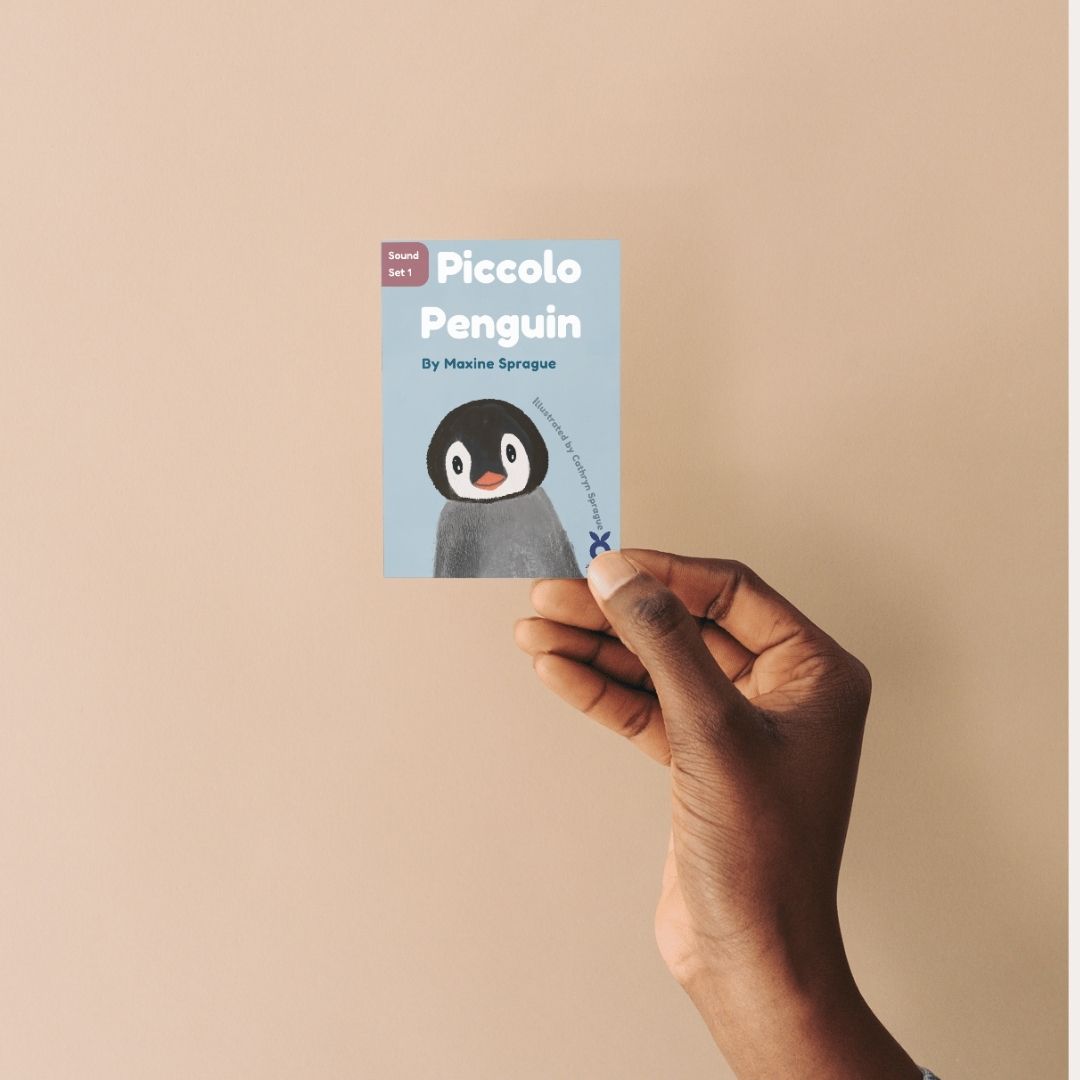 Hand holding a  mini children's book titled 'Piccolo Penguin' against a beige background